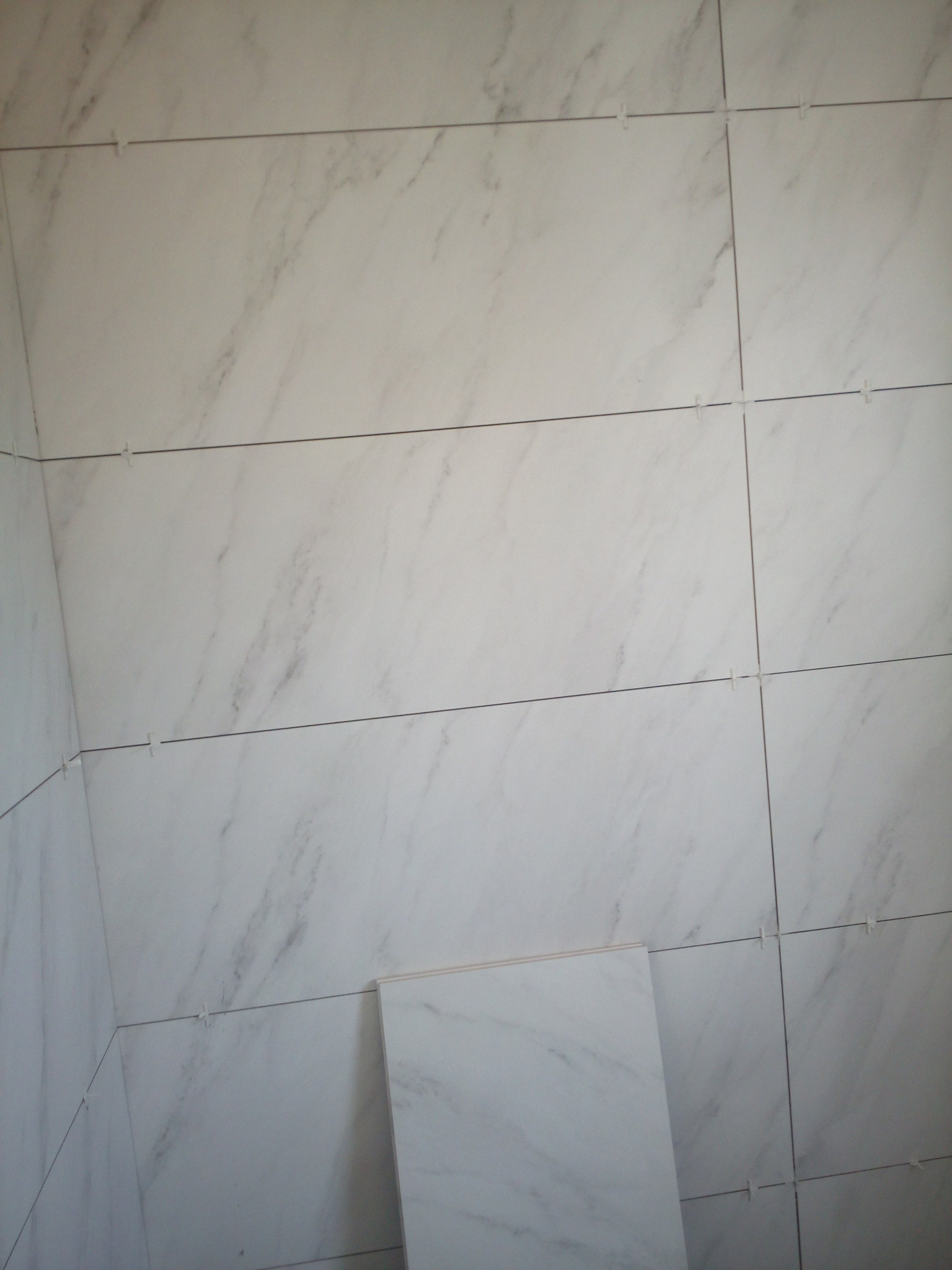 Ściana wyłożona białymi płytkami imitującymi marmur z krzyżykami dystansowymi, ułożona poziomo, z dodatkową płytką leżącą na podłodze.