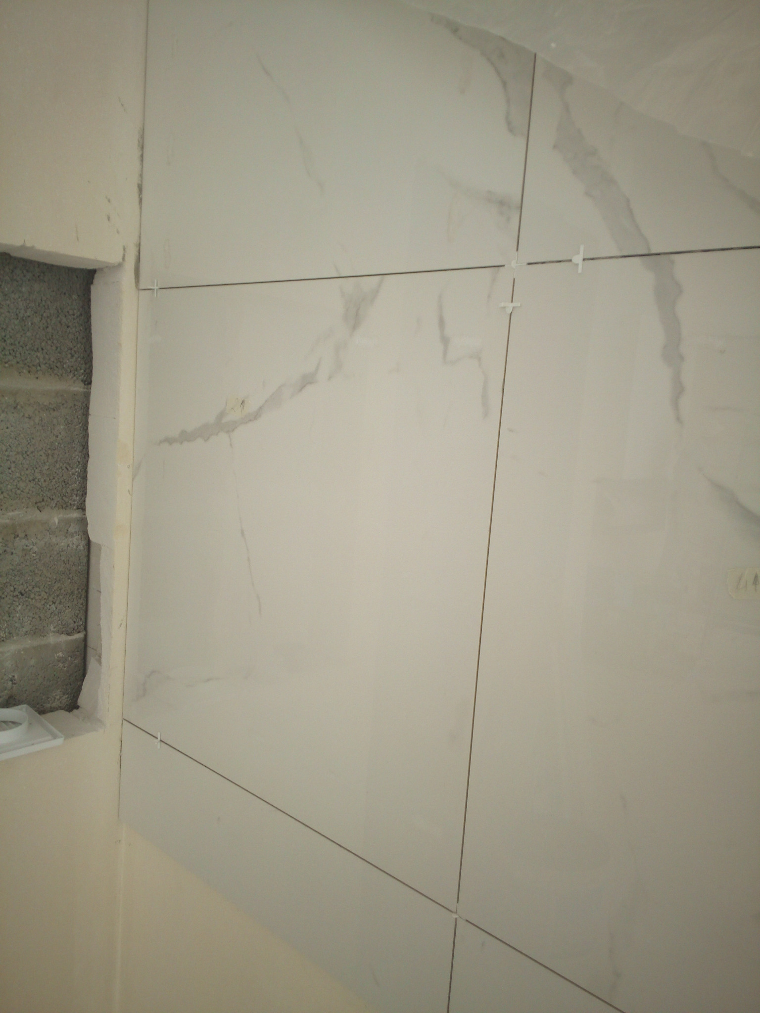 Układanie białych, marmurkowych płytek na ścianie w łazience, widoczne krzyżyki dystansowe i surowy mur z boku.