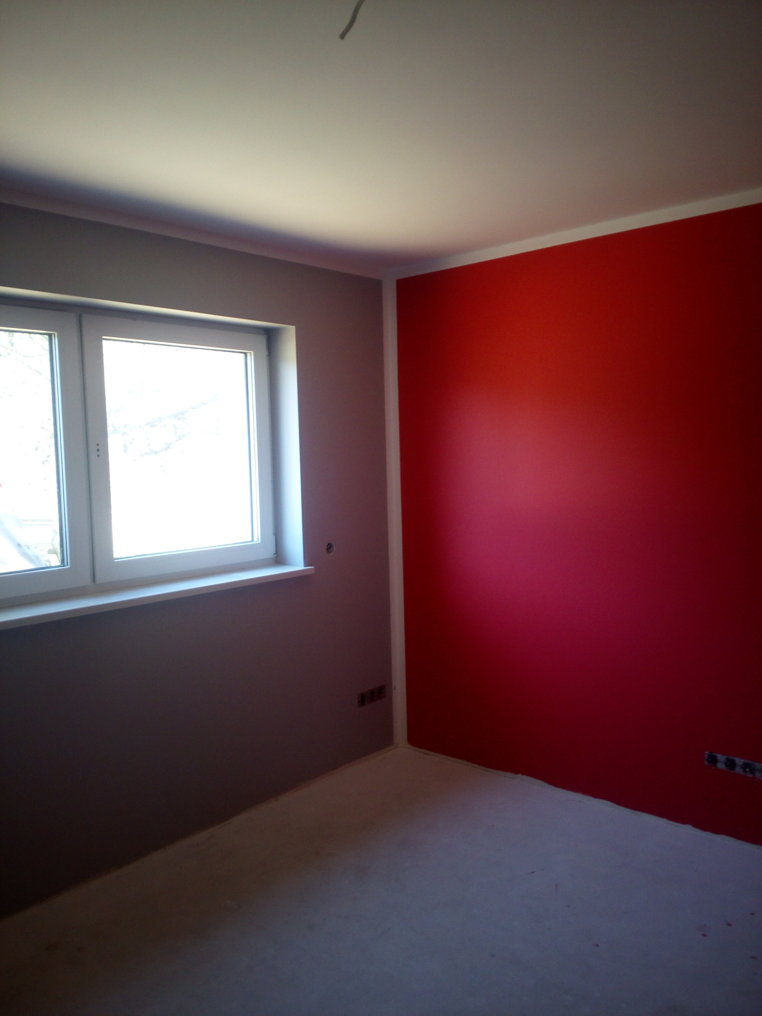 Pomieszczenie po malowaniu ścian: jedna ściana czerwona, druga szara, widoczne gniazdka elektryczne i białe okno.