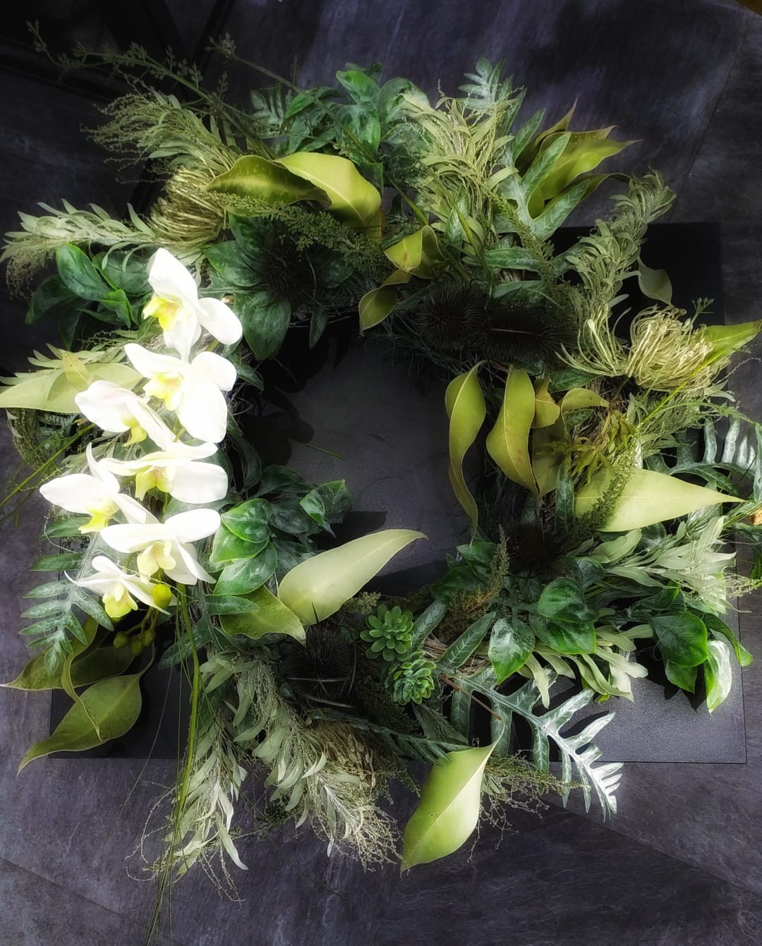 Dekoracyjny wieniec z białych orchidei i różnorodnych zielonych liści, ułożony na ciemnoszarym tle.