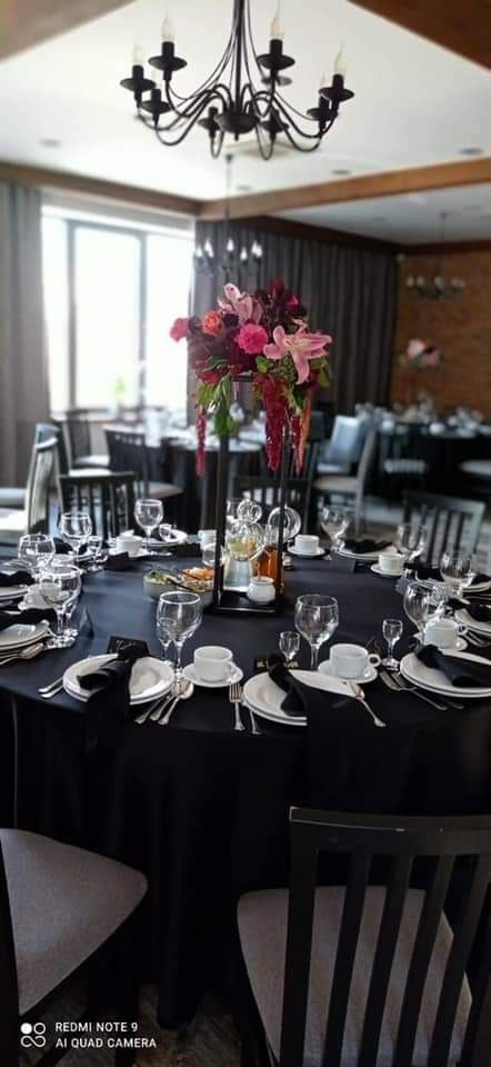 Elegancki stół bankietowy z czarnym obrusem, nakryty białą porcelaną, kryształowymi kieliszkami i czarnymi serwetkami. Centralny element stołu stanowi wysoka kompozycja kwiatowa w odcieniach różu...