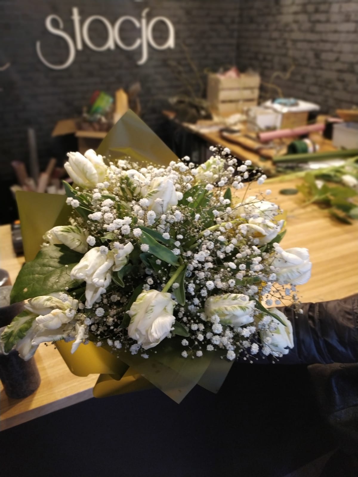 Biały bukiet z tulipanów i gipsówki w żółtym papierze, trzymany w ręku na tle drewnianego stołu i ściany z napisem 'Stacja'.