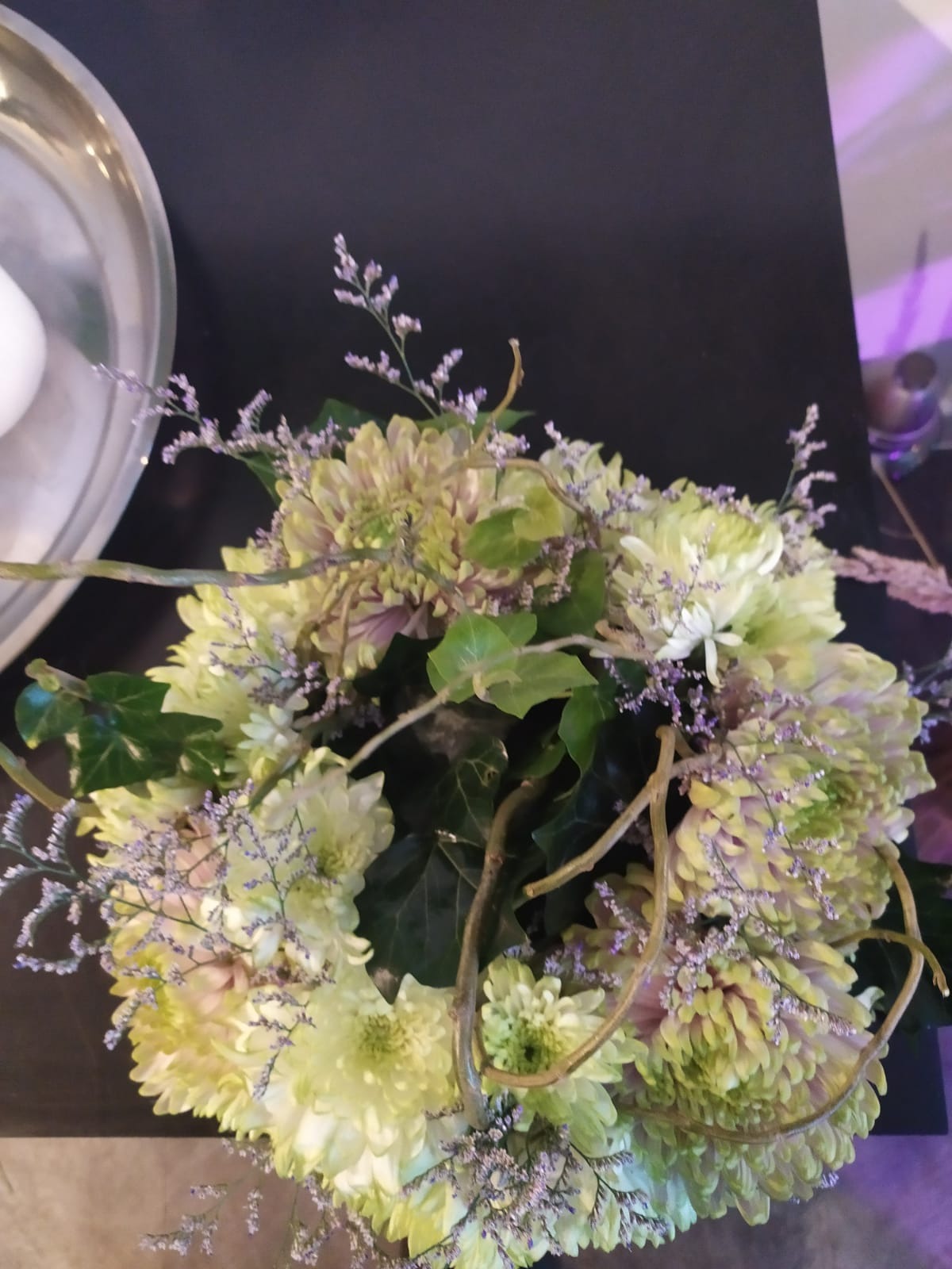 Kompozycja kwiatowa w odcieniach zieleni i fioletu z dodatkiem zielonych liści bluszczu i delikatnych gałązek, ułożona na ciemnym tle, obok srebrnej tacy.