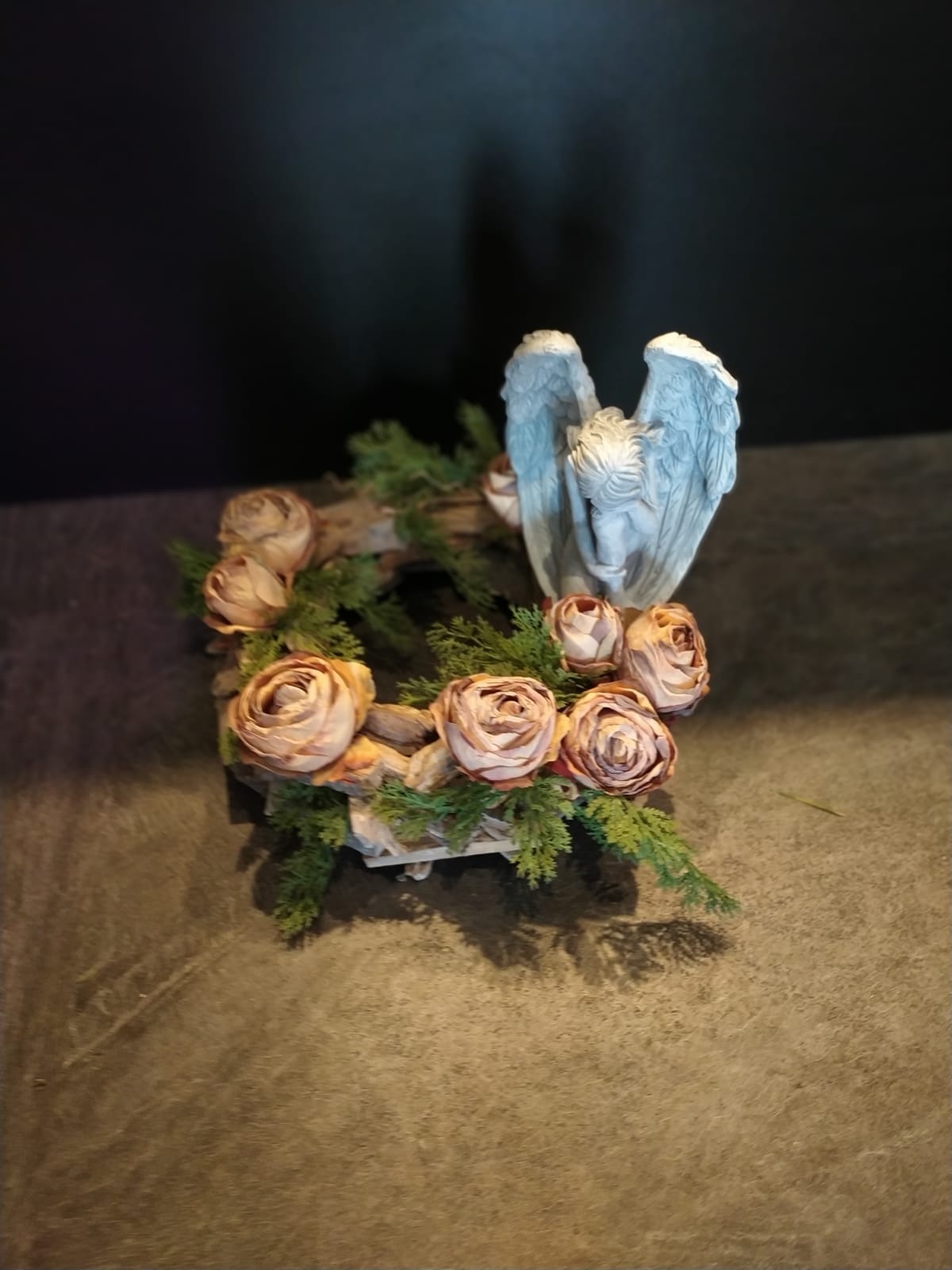 Kompozycja dekoracyjna: wieniec z suszonych róż i zielonych gałązek, z figurką anioła z pochyloną głową, na ciemnym tle.