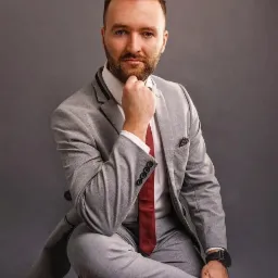 Portret mężczyzny w szarym garniturze z czerwoną wstążką, siedzącego na krześle na tle szarego tła, z ręką podpierającą brodę i zegarkiem na nadgarstku.