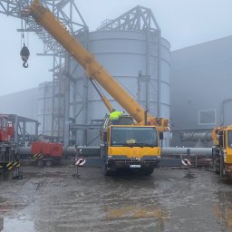 KRIS Dźwigi Lubin, Usługi dźwigowe, Wynajem dźwigów - Trzy dźwigi samochodowe, czerwony i dwa żółte, ustawione na placu budowy w mglisty dzień, z widocznymi silosami w tle.