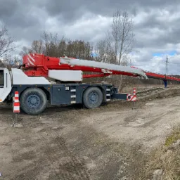 Biało-czerwony dźwig samochodowy ustawiony na utwardzonym, szutrowym podłożu z wysuniętym ramieniem w kierunku pracującego operatora, na tle pochmurnego nieba i linii wysokiego napięcia.