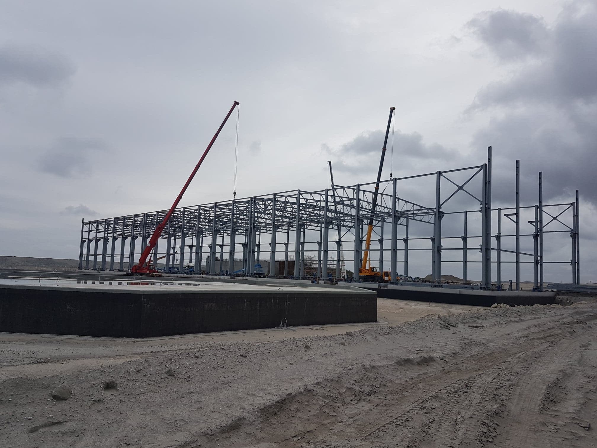 Montaż konstrukcji stalowej hali przemysłowej z użyciem dwóch żurawi na placu budowy w pochmurny dzień.