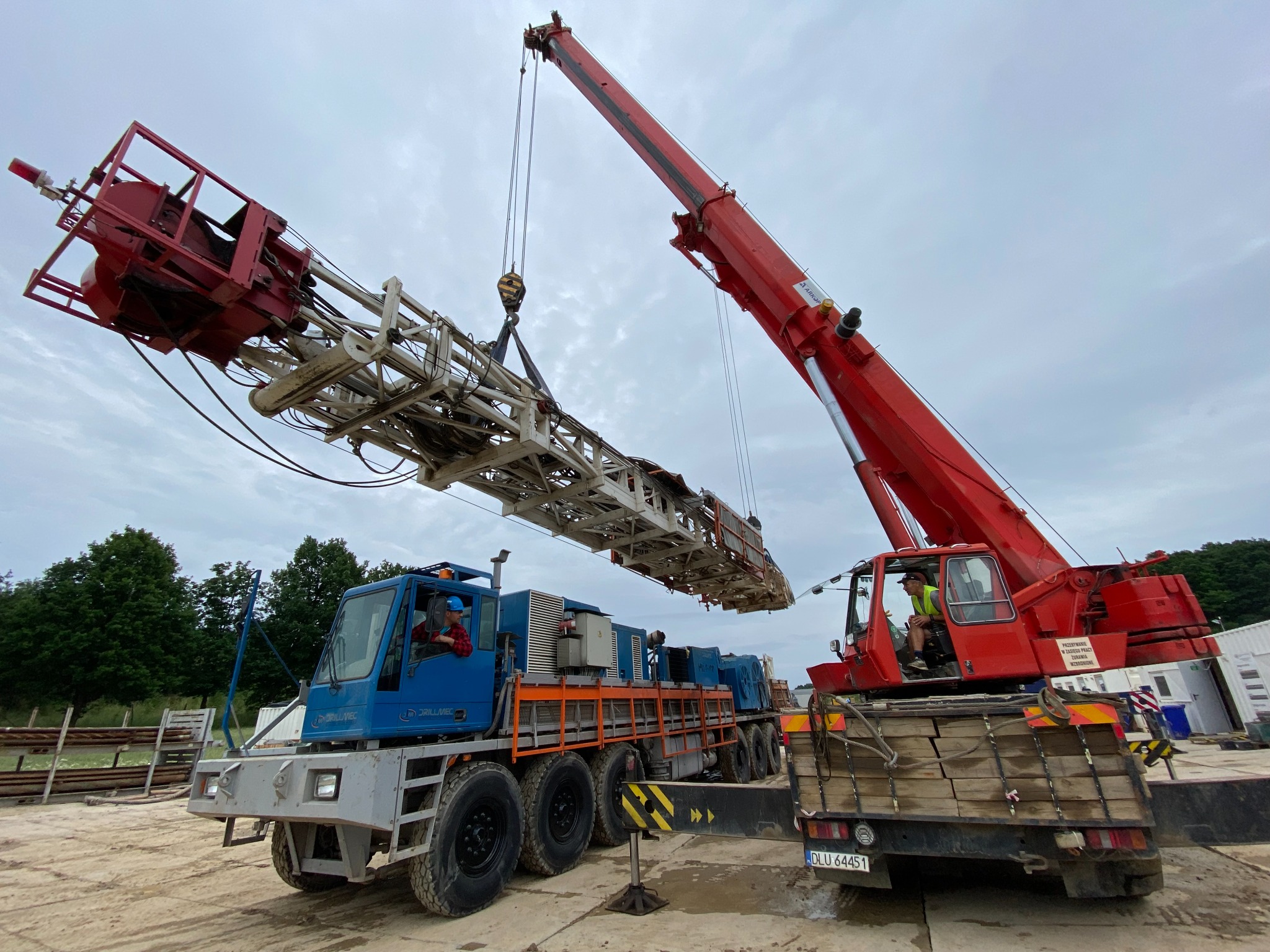 Czerwony dźwig podnosi element konstrukcyjny na tle pochmurnego nieba, z widocznym operatorem w kabinie i niebieską ciężarówką Drillmec z operatorem w tle.