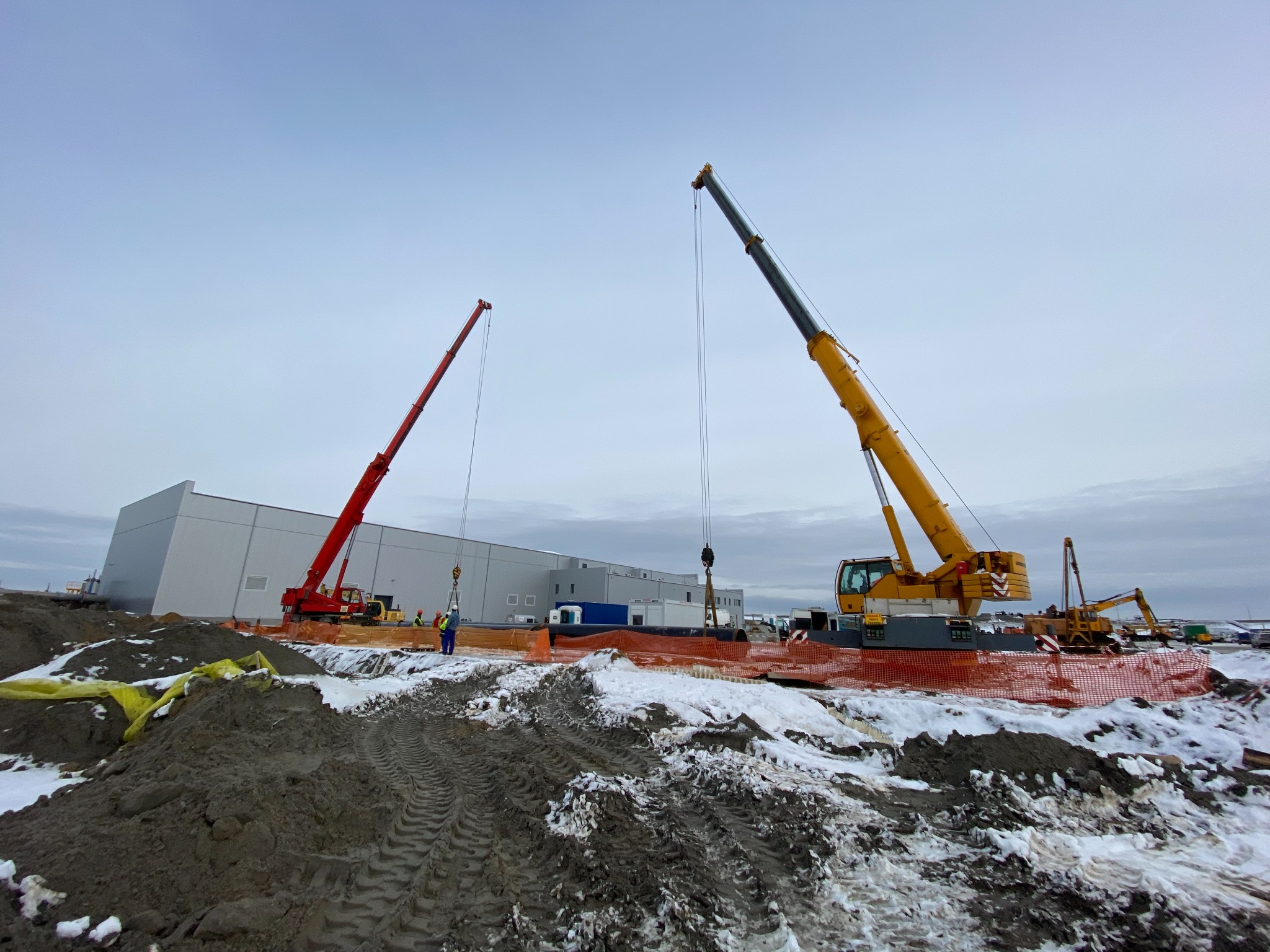 Dwa dźwigi: czerwony i żółty, unoszą rury na placu budowy nowego magazynu w pochmurny dzień. W tle widoczny biały budynek magazynowy.