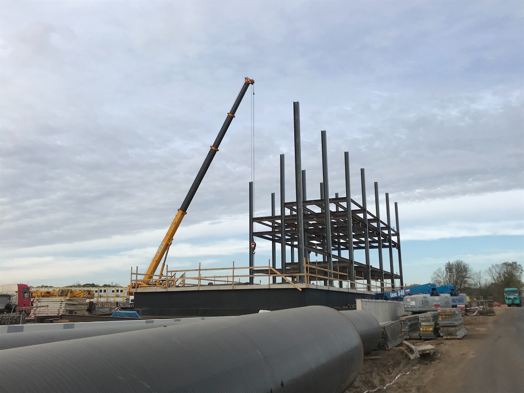 Żółty dźwig teleskopowy w trakcie montażu stalowej konstrukcji budynku, widoczne rury na pierwszym planie i pochmurne niebo w tle.