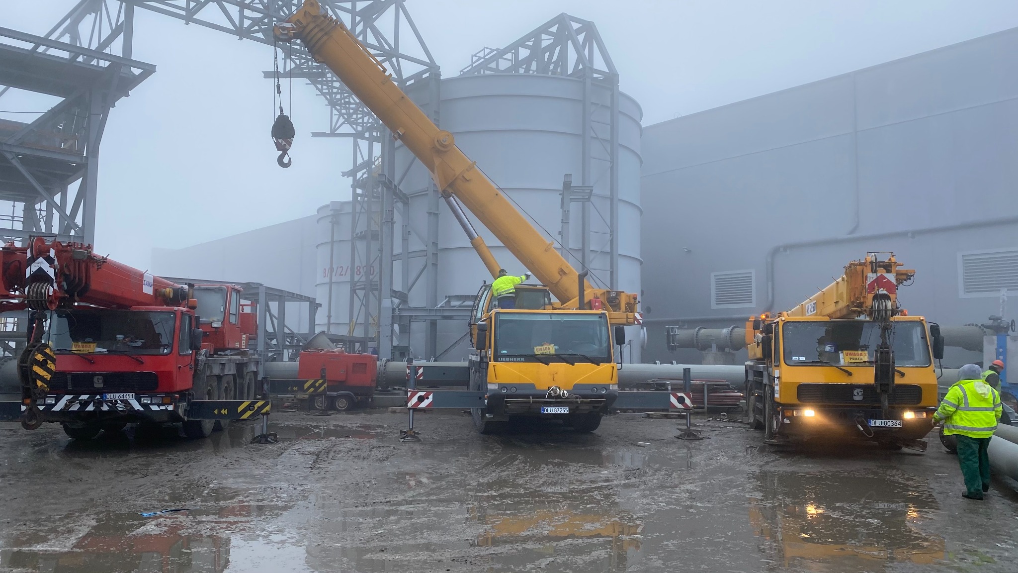 Trzy dźwigi samochodowe, czerwony i dwa żółte, ustawione na placu budowy w mglisty dzień, z widocznymi silosami w tle.