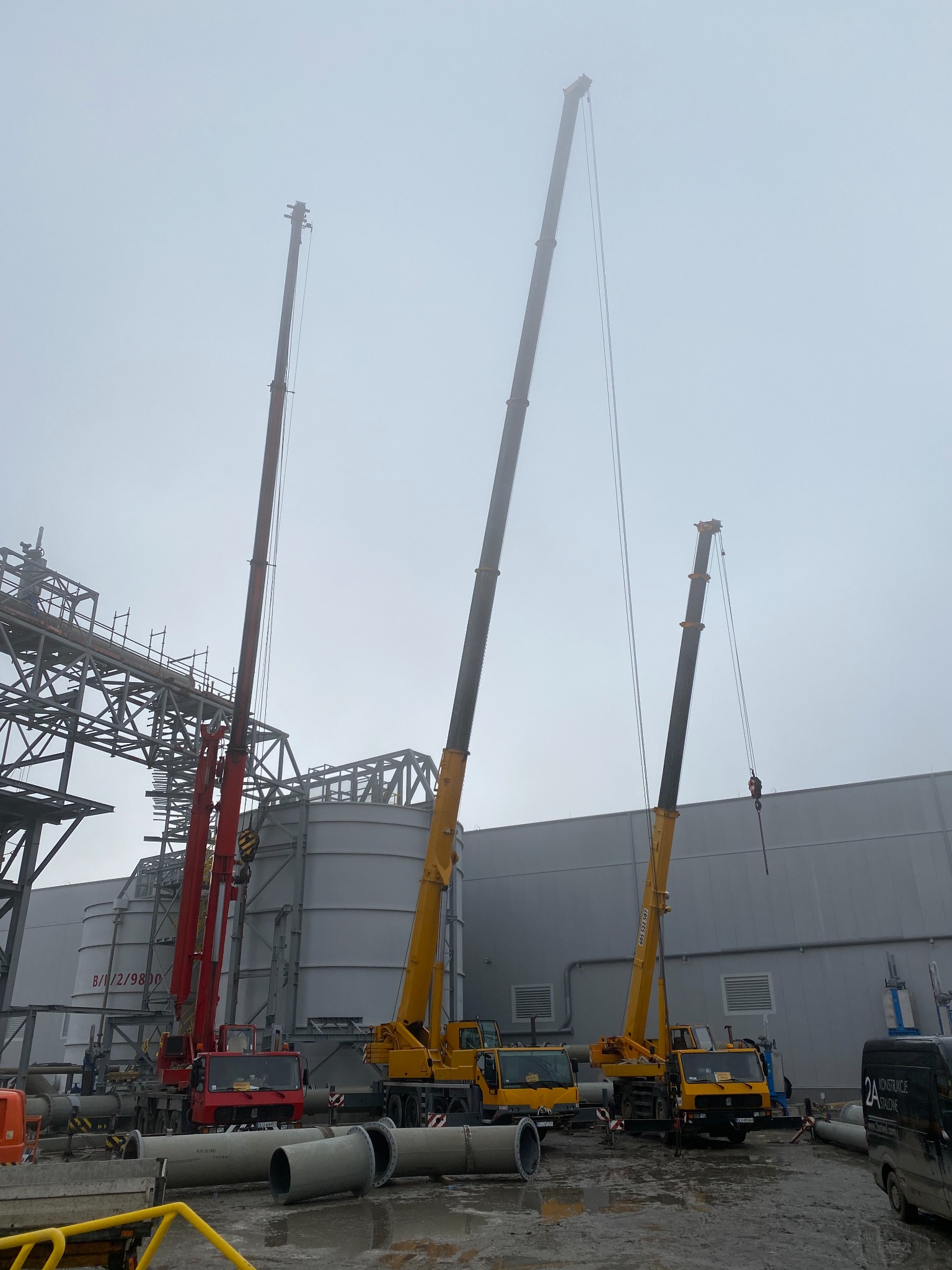 Trzy dźwigi (dwa żółte i jeden czerwony) na placu budowy w Lubinie, w tle silosy i konstrukcja stalowa, pochmurny dzień.