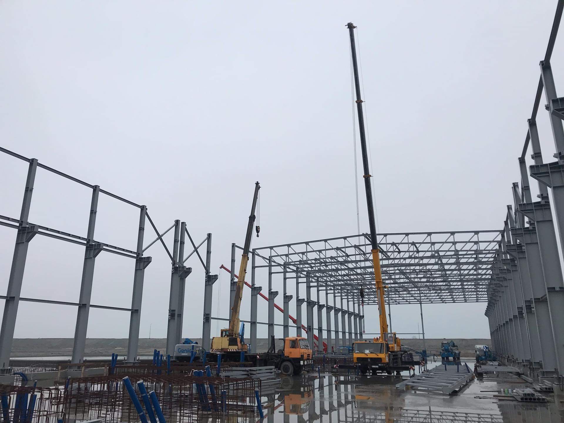 Montaż stalowej konstrukcji hali przemysłowej przy użyciu dwóch dźwigów samochodowych w pochmurny dzień.