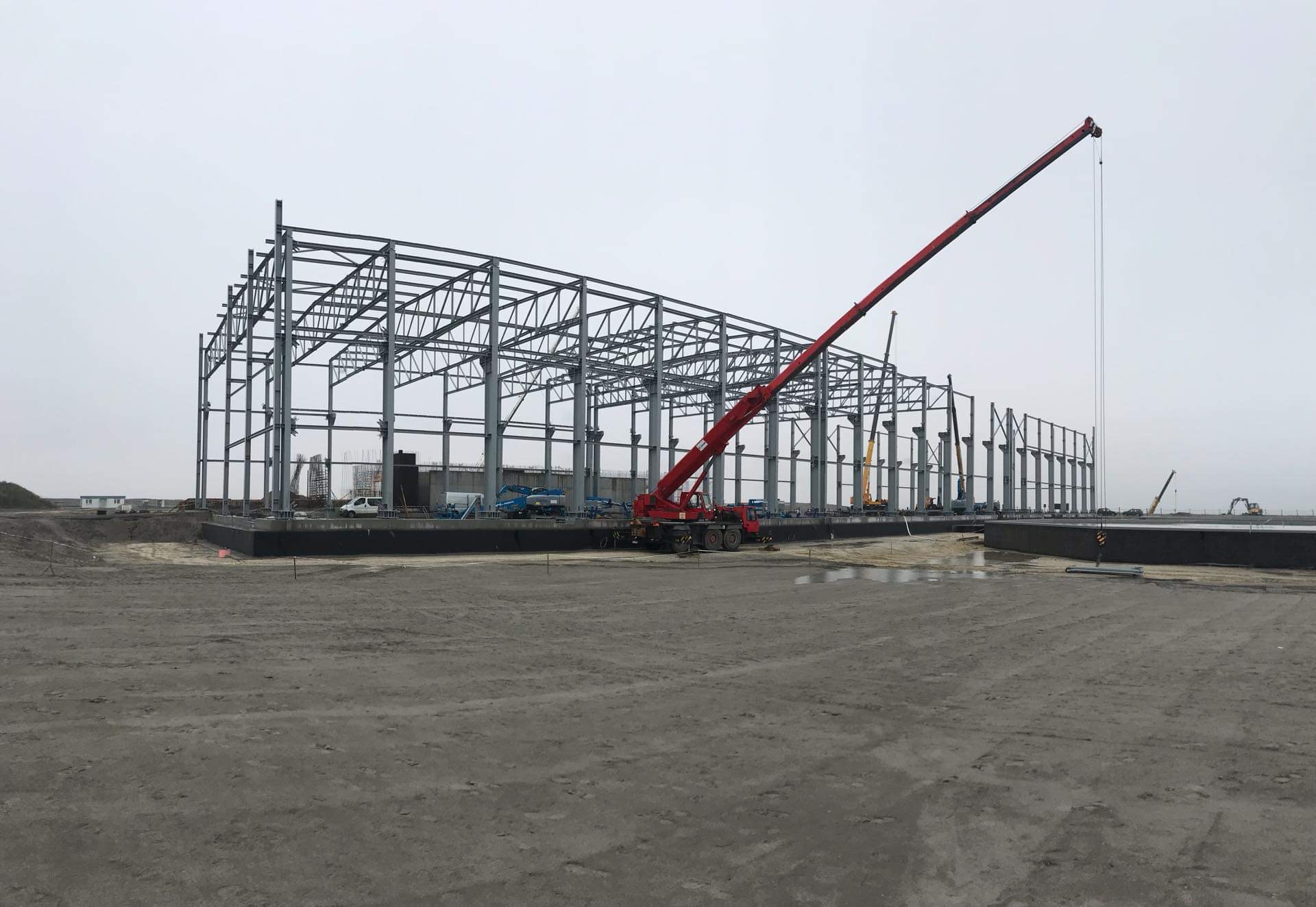 Czerwony dźwig na budowie hali stalowej, widoczne elementy konstrukcyjne, teren przygotowany pod wylewkę fundamentów.