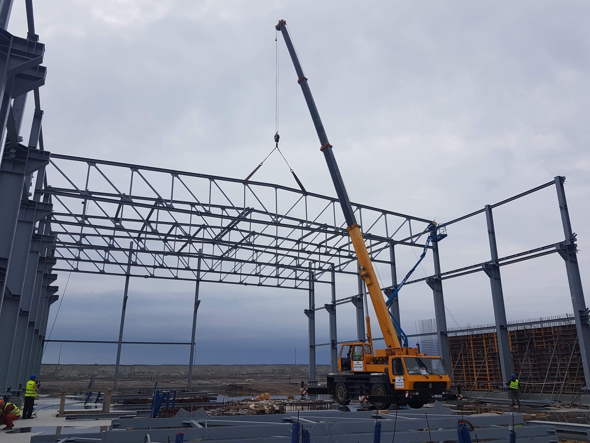 Żółty dźwig samochodowy podnosi element stalowej konstrukcji szkieletu hali przemysłowej na tle pochmurnego nieba.