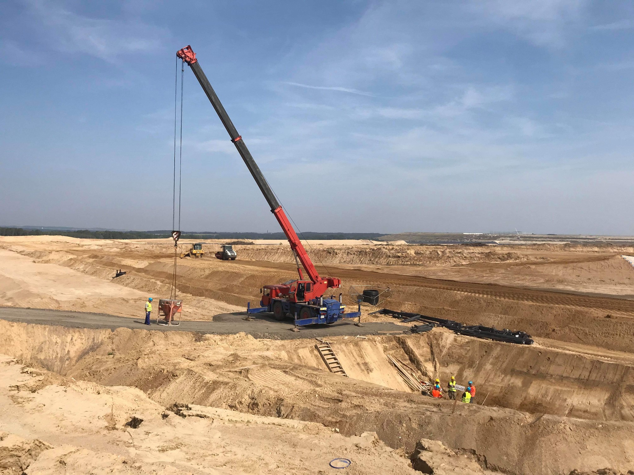 Czerwono-niebieski dźwig samochodowy na terenie budowy z piaskiem, podnosi ładunek nad wykopem, w tle widoczne maszyny budowlane i pracownicy w kaskach.