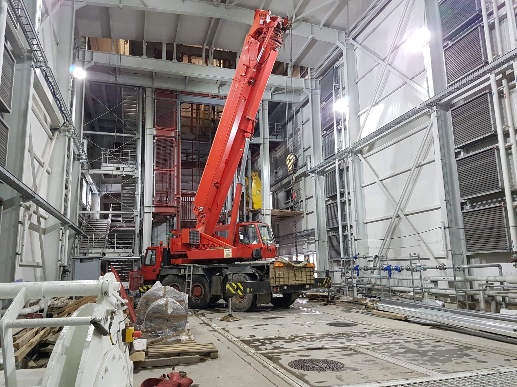 Czerwony żuraw samojezdny z wysuniętym ramieniem wewnątrz hali przemysłowej o konstrukcji stalowej, widoczne elementy instalacji i materiały budowlane.