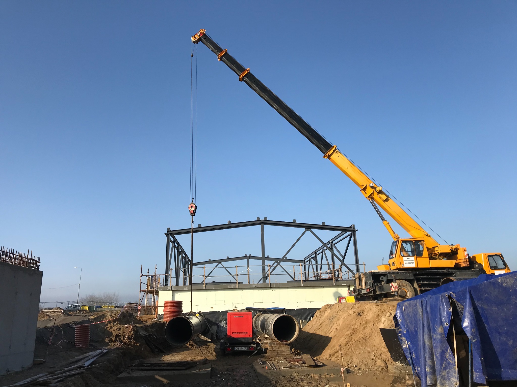Żółty dźwig teleskopowy podnosi element konstrukcji stalowej na placu budowy, widoczne rury kanalizacyjne, generator prądu i fragmenty betonowych ścian.