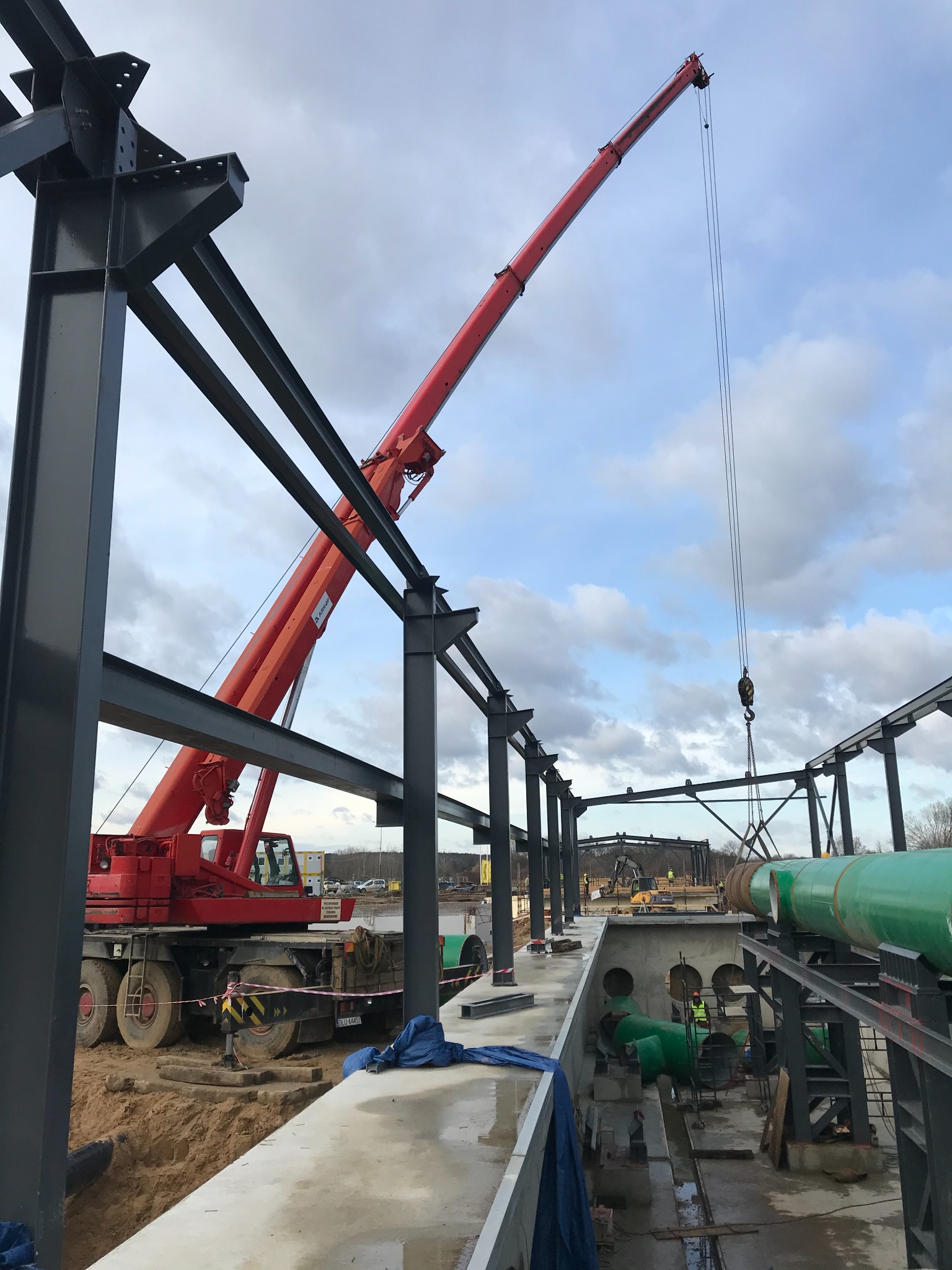 Czerwony dźwig teleskopowy podnosi rury na placu budowy, widoczne stalowe konstrukcje i pracownicy w odblaskowych kamizelkach.