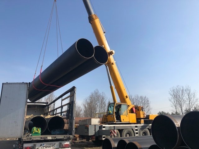 Żółty dźwig samochodowy z wysięgnikiem podnosi dwie długie, czarne rury i umieszcza je na skrzyni samochodu ciężarowego marki Schmitz, na tle błękitnego nieba i drzew bez liści.