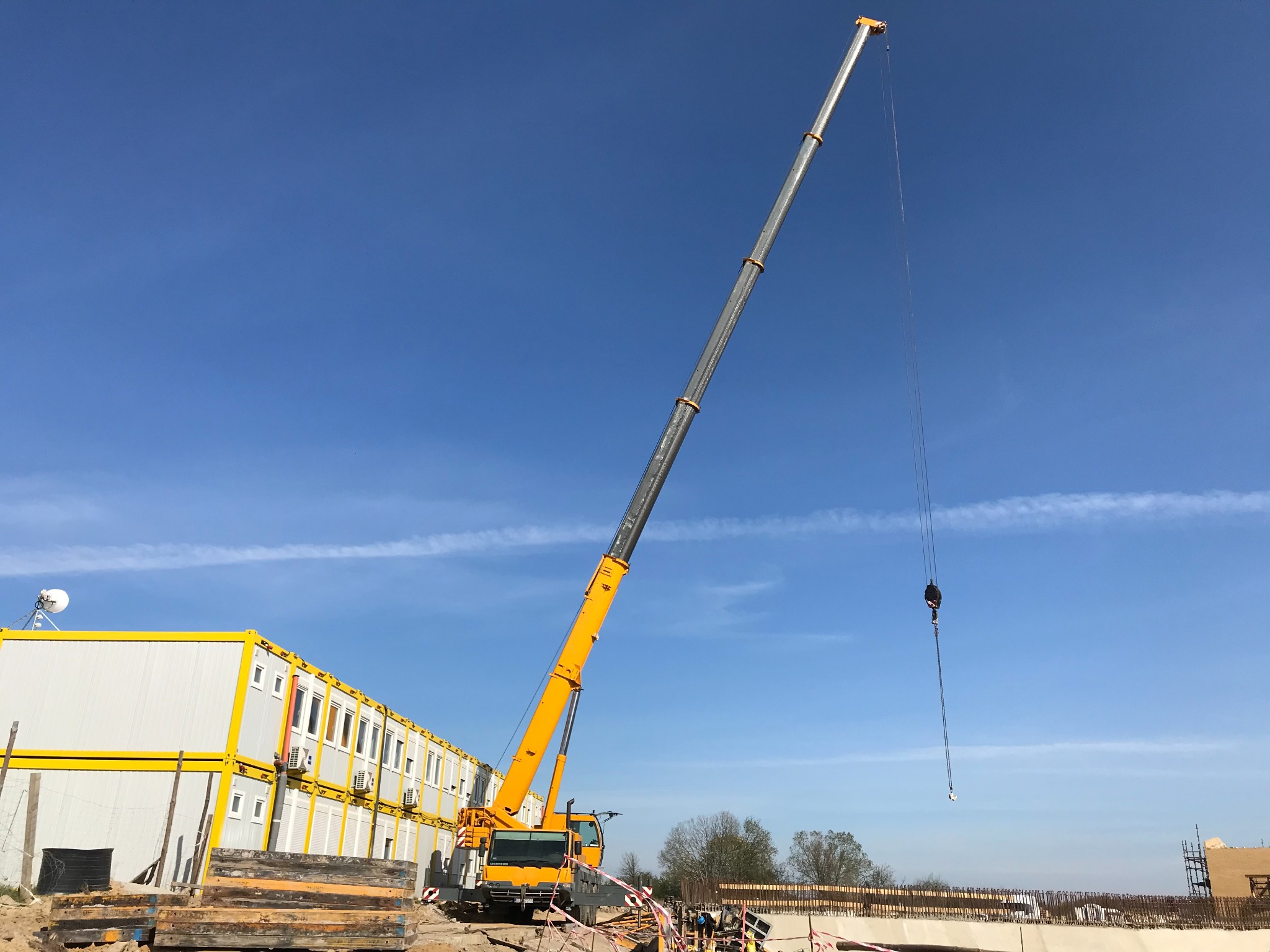 Żółty dźwig teleskopowy Liebherr na placu budowy z kontenerami biurowymi w tle, błękitne niebo z smugami kondensacyjnymi.