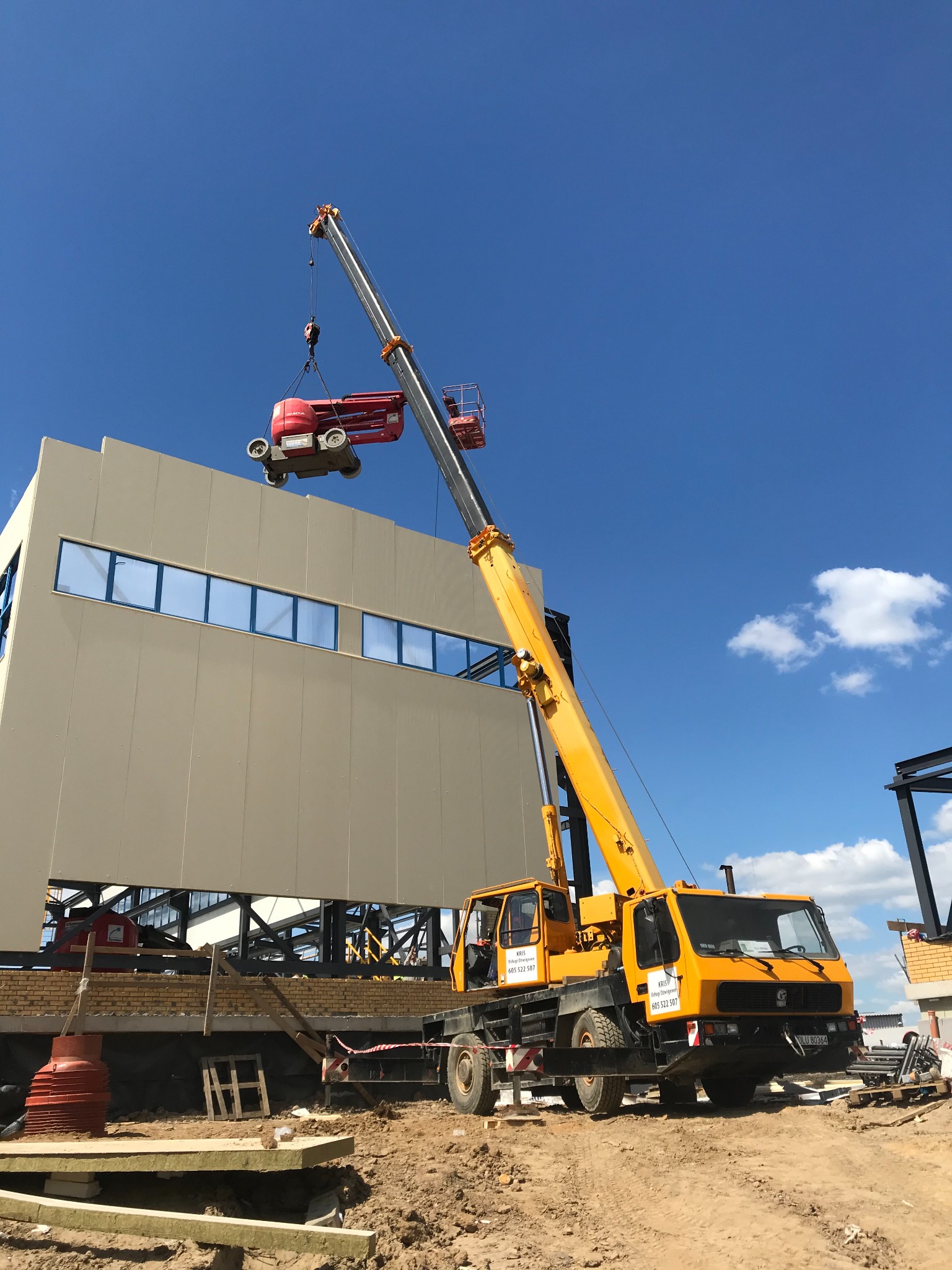 Żółty dźwig samochodowy podnosi czerwoną platformę roboczą na tle jasnej ściany budynku z niebieskimi oknami i błękitnego nieba.