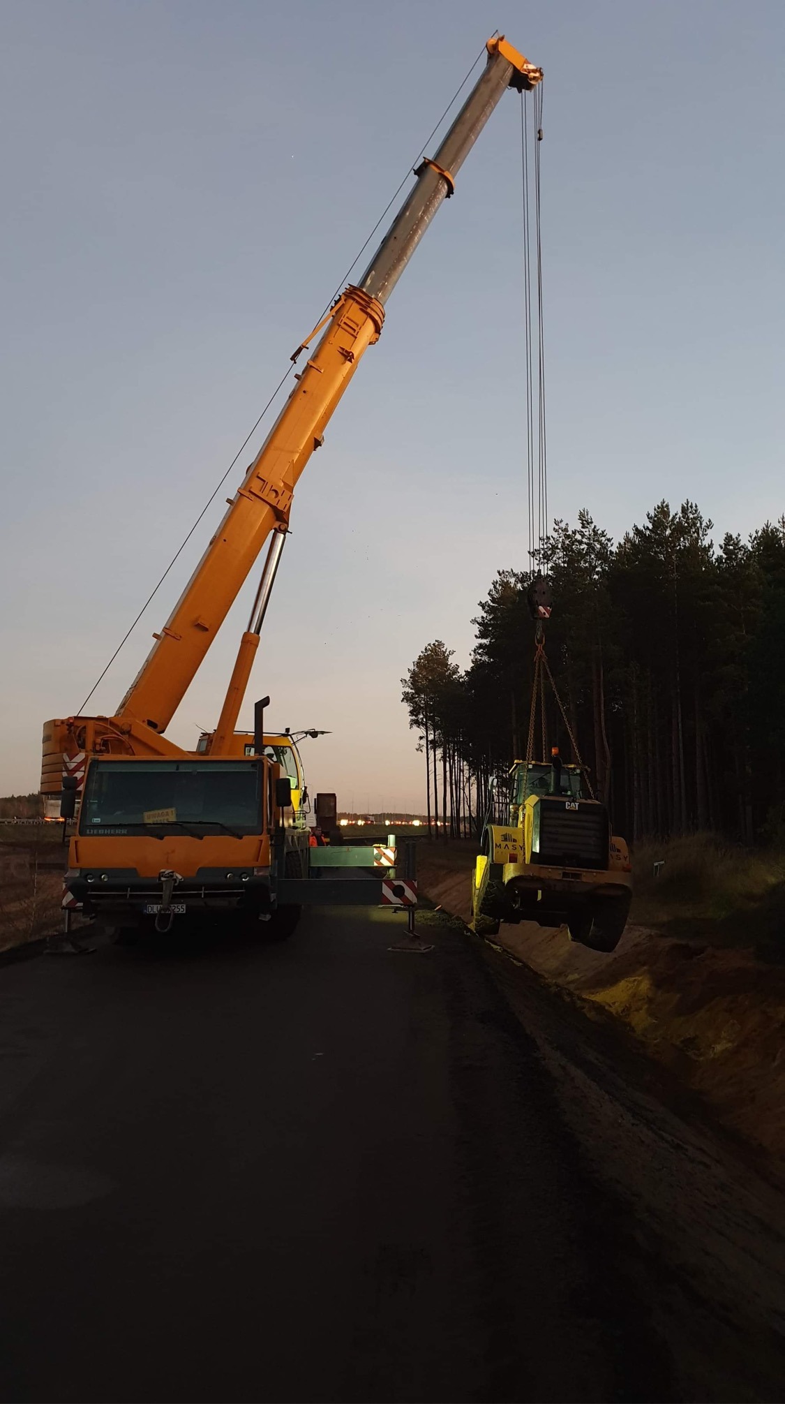 Żółty dźwig Liebherr podnosi żółtą ładowarkę CAT na tle lasu i zachodzącego słońca.