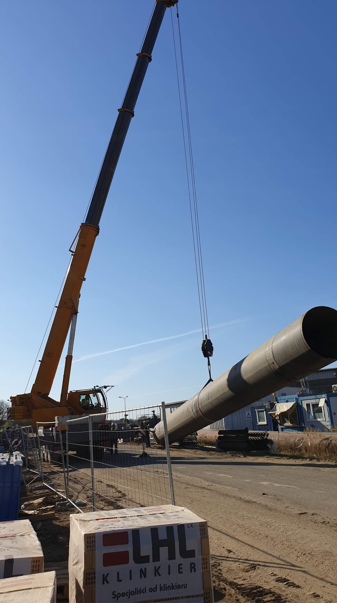 Żółty dźwig podnosi betonową rurę na tle błękitnego nieba, w tle widoczni pracownicy budowlani oraz kontenery biurowe, na pierwszym planie paleta z cegłami klinkierowymi.