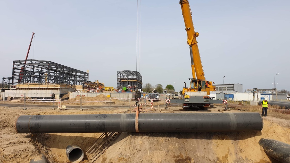 Żółty dźwig podnosi rurę kanalizacyjną na placu budowy, w tle stalowa konstrukcja budynku i pracownik w kamizelce odblaskowej.