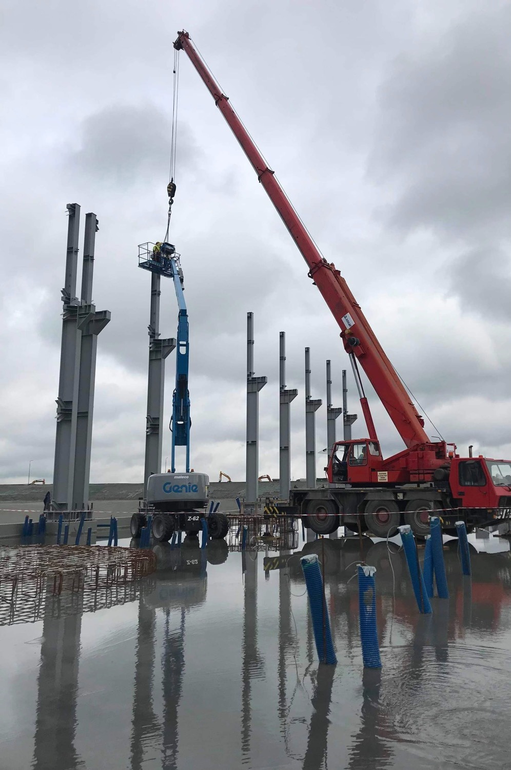 Montaż stalowych słupów konstrukcyjnych na mokrym placu budowy przy użyciu czerwonego dźwigu samochodowego i niebieskiego podnośnika koszowego Genie Z-62.