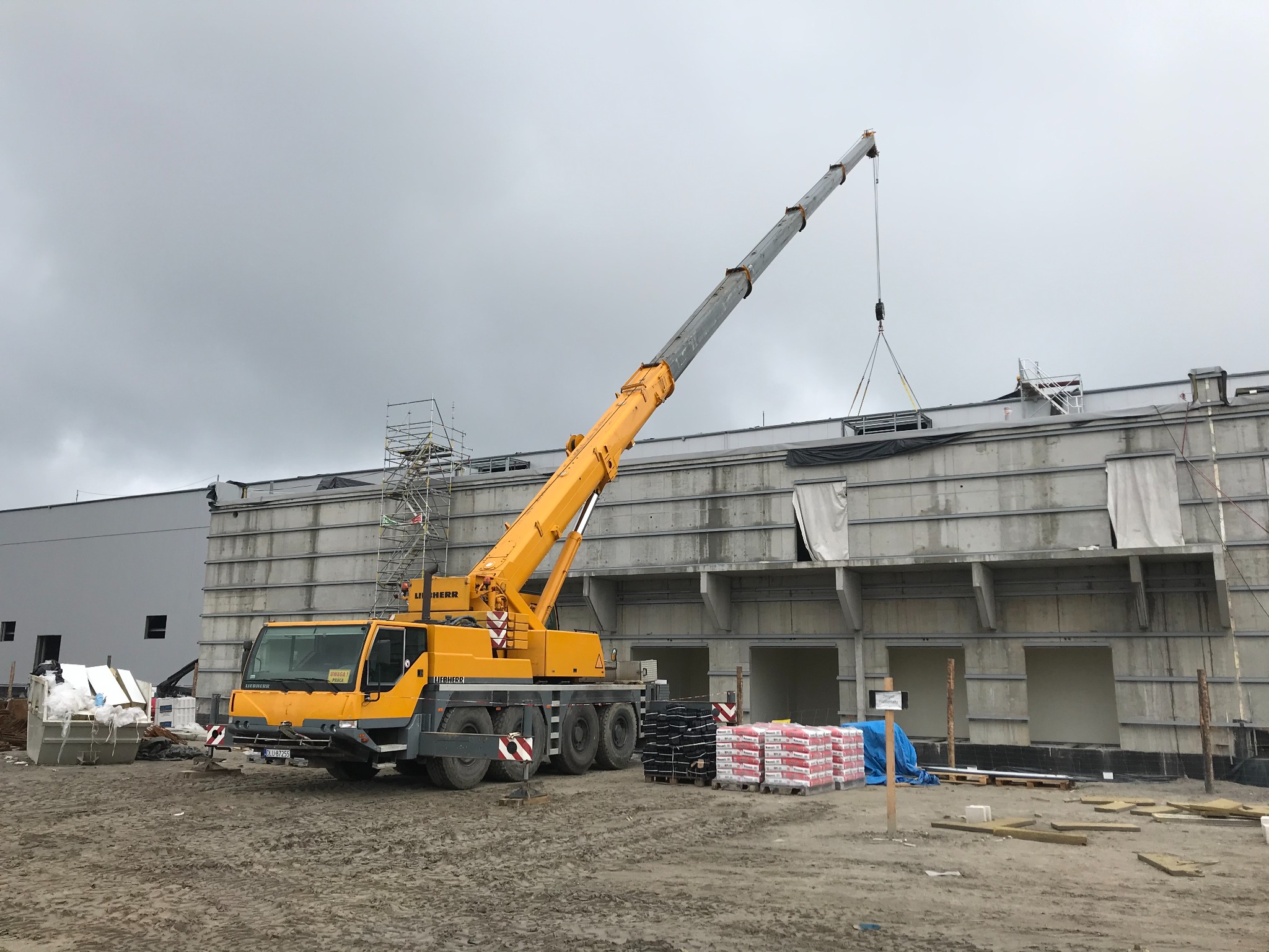Żółty dźwig Liebherr podnosi element konstrukcyjny na budowę hali w pochmurny dzień, widoczne rusztowania i materiały budowlane.
