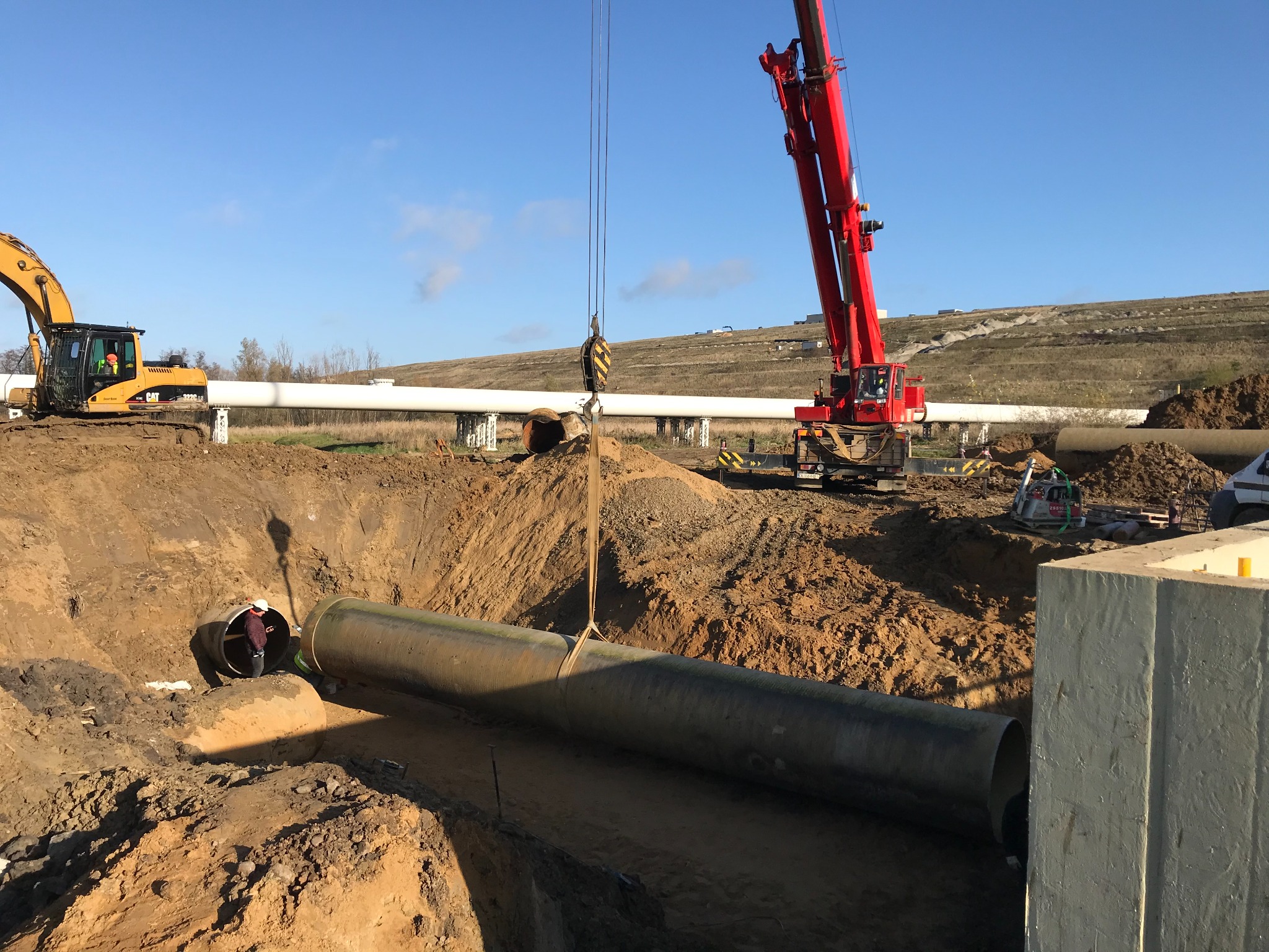 Czerwony dźwig opuszcza rurę do wykopu, w tle widoczny pracownik w kasku, żółta koparka i biały rurociąg na podporach.