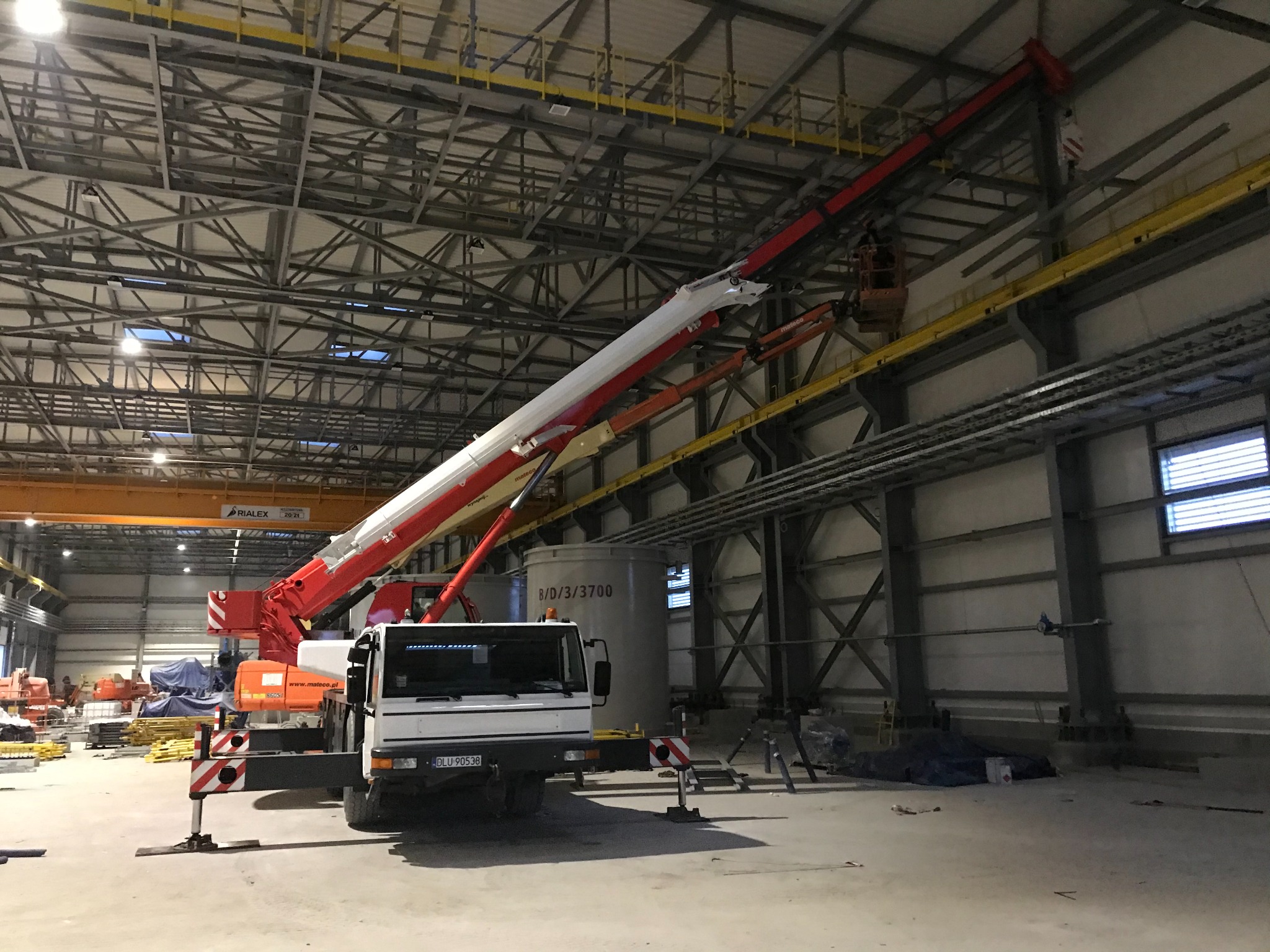 Wnętrze hali przemysłowej z widocznym dźwigiem samochodowym z podnośnikiem koszowym, pracownik w koszu na wysokości przy konstrukcji dachu, w tle inne maszyny i materiały budowlane.
