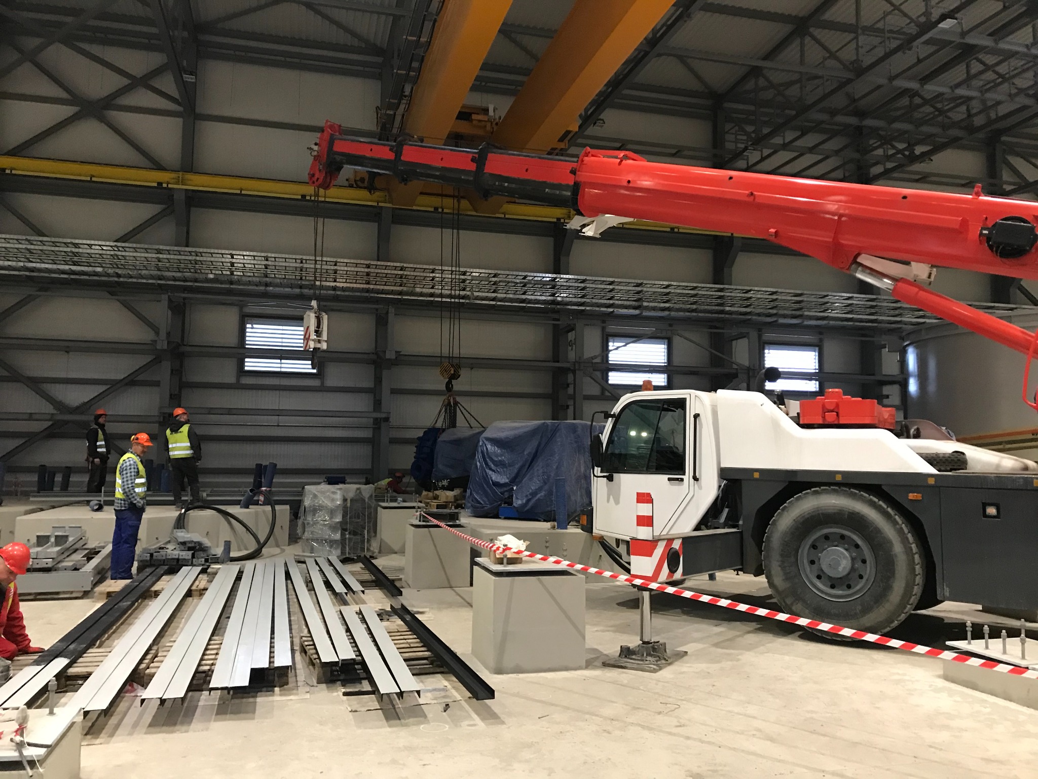 Wnętrze hali produkcyjnej z suwnicą podnoszącą element pod plandeką, pracownicy w odblaskowych kamizelkach nadzorują operację, na pierwszym planie leżą metalowe profile.