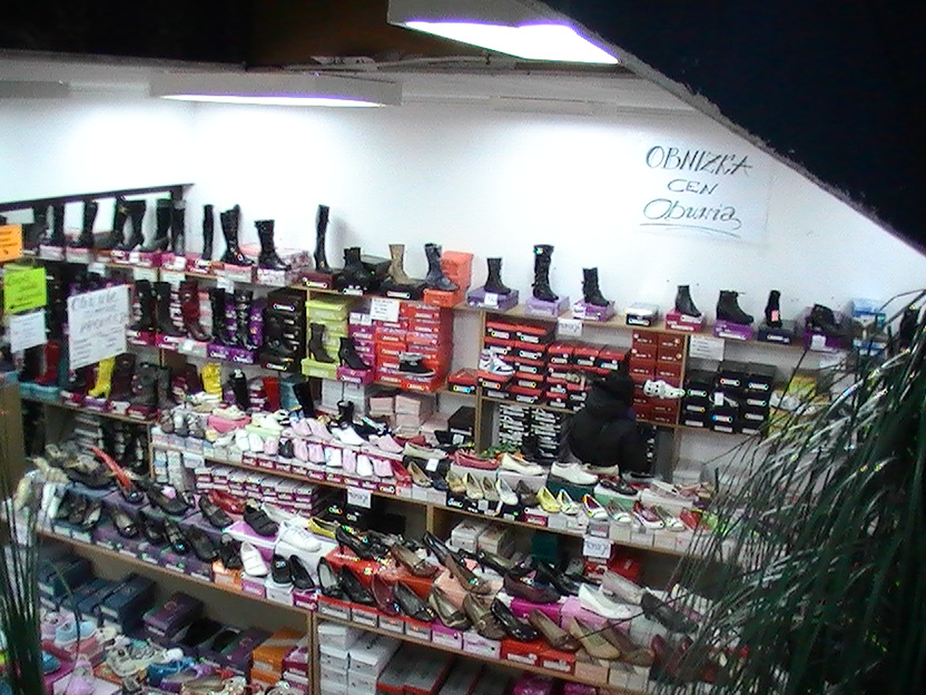 Wnętrze sklepu obuwniczego z ekspozycją różnorodnych modeli butów damskich, męskich i dziecięcych na półkach, widoczny napis 'Obniżka cen Obuwia' na ścianie.