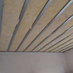 Montaż izolacji z wełny mineralnej w metalowym stelażu sufitu podwieszanego w jasnym pomieszczeniu z oknem.