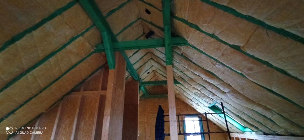 Ocieplenie poddasza wełną mineralną, widoczne drewniane krokwie pomalowane na zielono, fragment okna w tle.