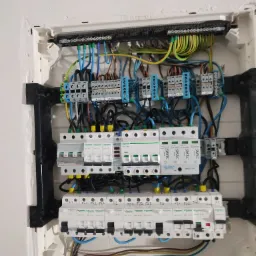 Otwarta skrzynka rozdzielcza z widocznymi bezpiecznikami, przewodami i listwami zaciskowymi, zamontowana na białej ścianie z widocznymi niedoskonałościami wykończenia.