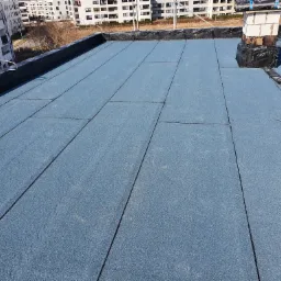 Niebieska papa termozgrzewalna na płaskim dachu budynku wielorodzinnego, widoczny komin z cegieł i niezabezpieczone rolki papy, w tle nowoczesna architektura.