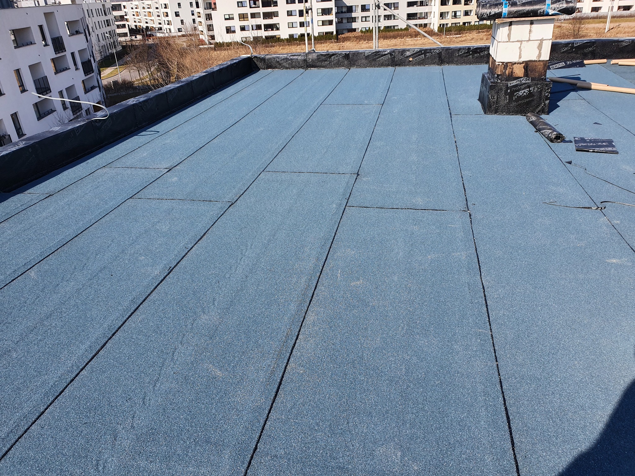 Niebieska papa termozgrzewalna na płaskim dachu budynku wielorodzinnego, widoczny komin z cegieł i niezabezpieczone rolki papy, w tle nowoczesna architektura.