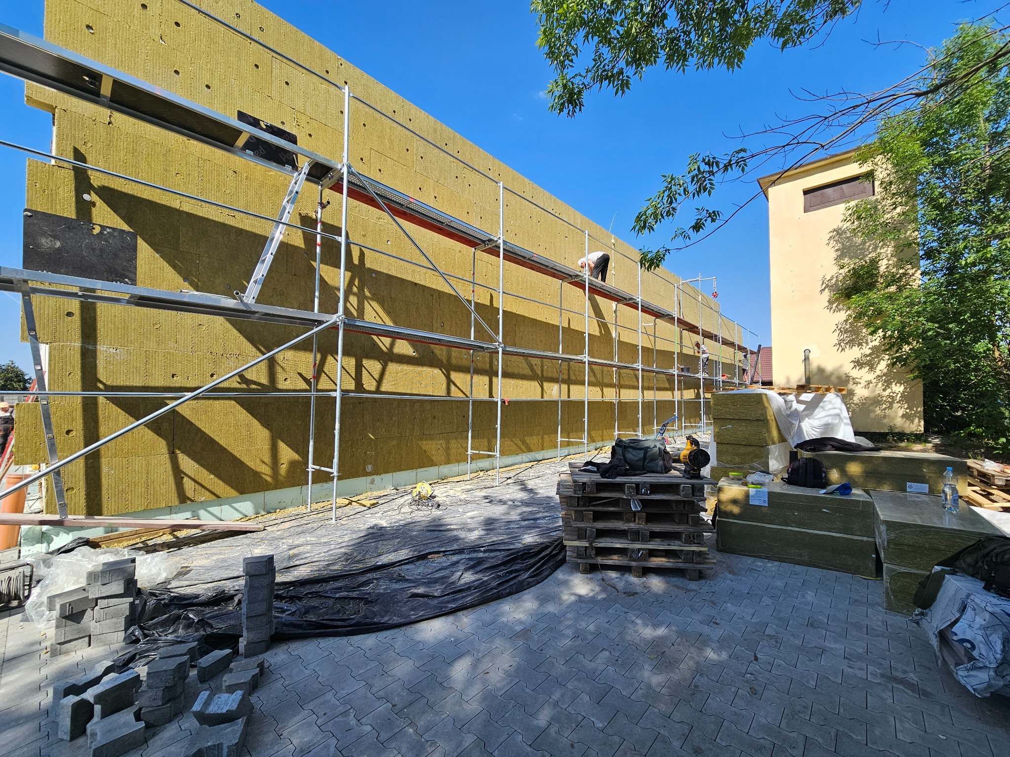 Ocieplanie elewacji budynku wełną mineralną. Widoczne rusztowania, materiały izolacyjne i pracownik na rusztowaniu. Prace w toku, słoneczny dzień.