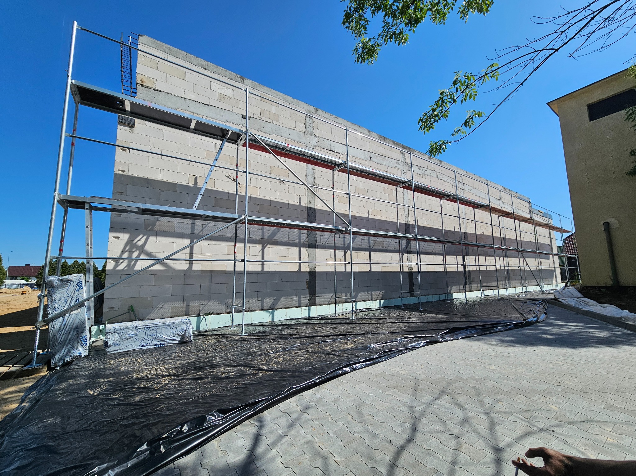 Ściana budynku w trakcie budowy z białych bloczków, otoczona rusztowaniem, widok z zewnątrz w słoneczny dzień. Widoczna izolacja fundamentów.