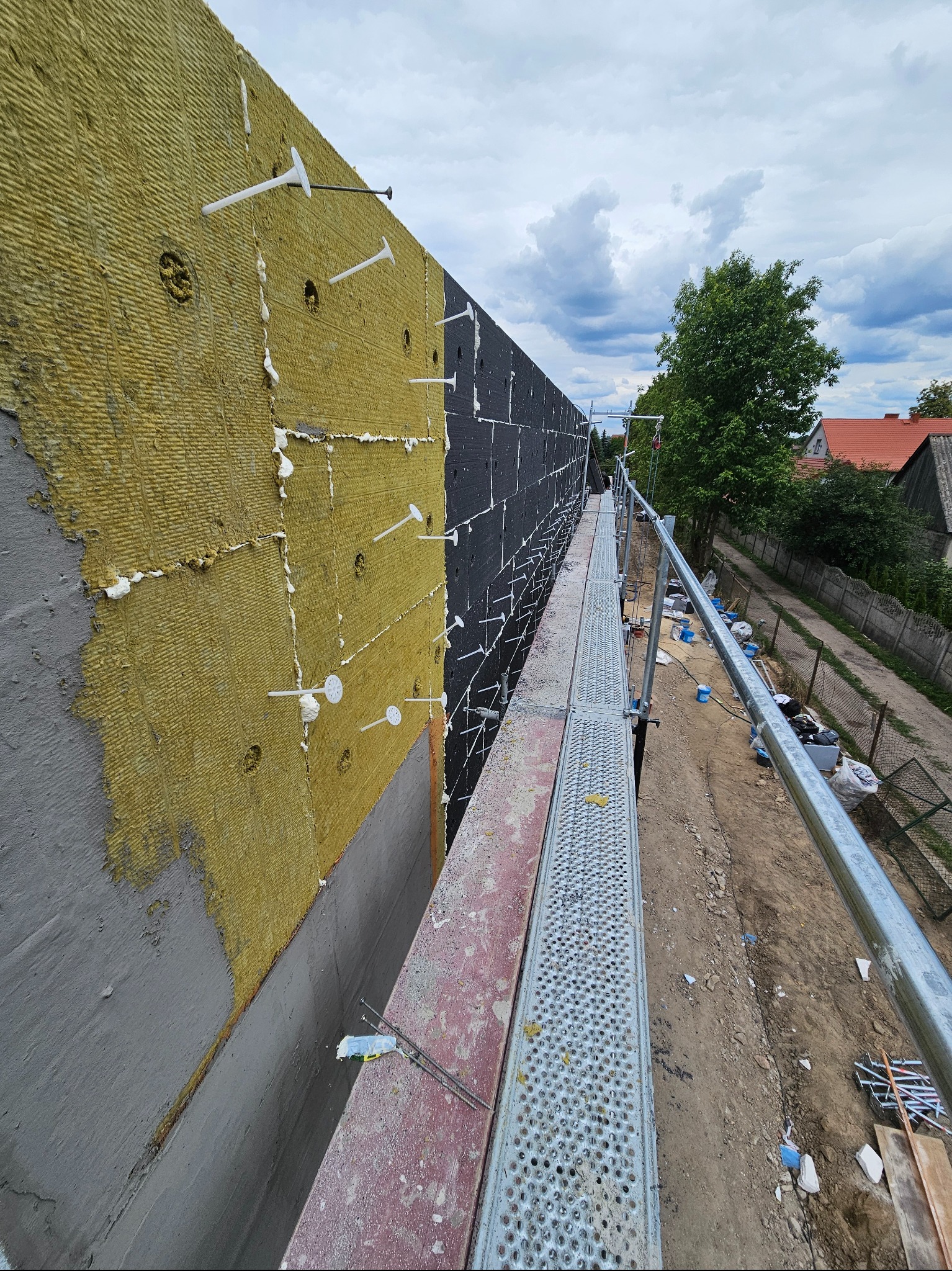 Ocieplanie ściany budynku wełną mineralną, widoczne kołki mocujące i rusztowanie. Prace na wysokości, etap ocieplania. Tło: zabudowa jednorodzinna i pochmurne niebo.
