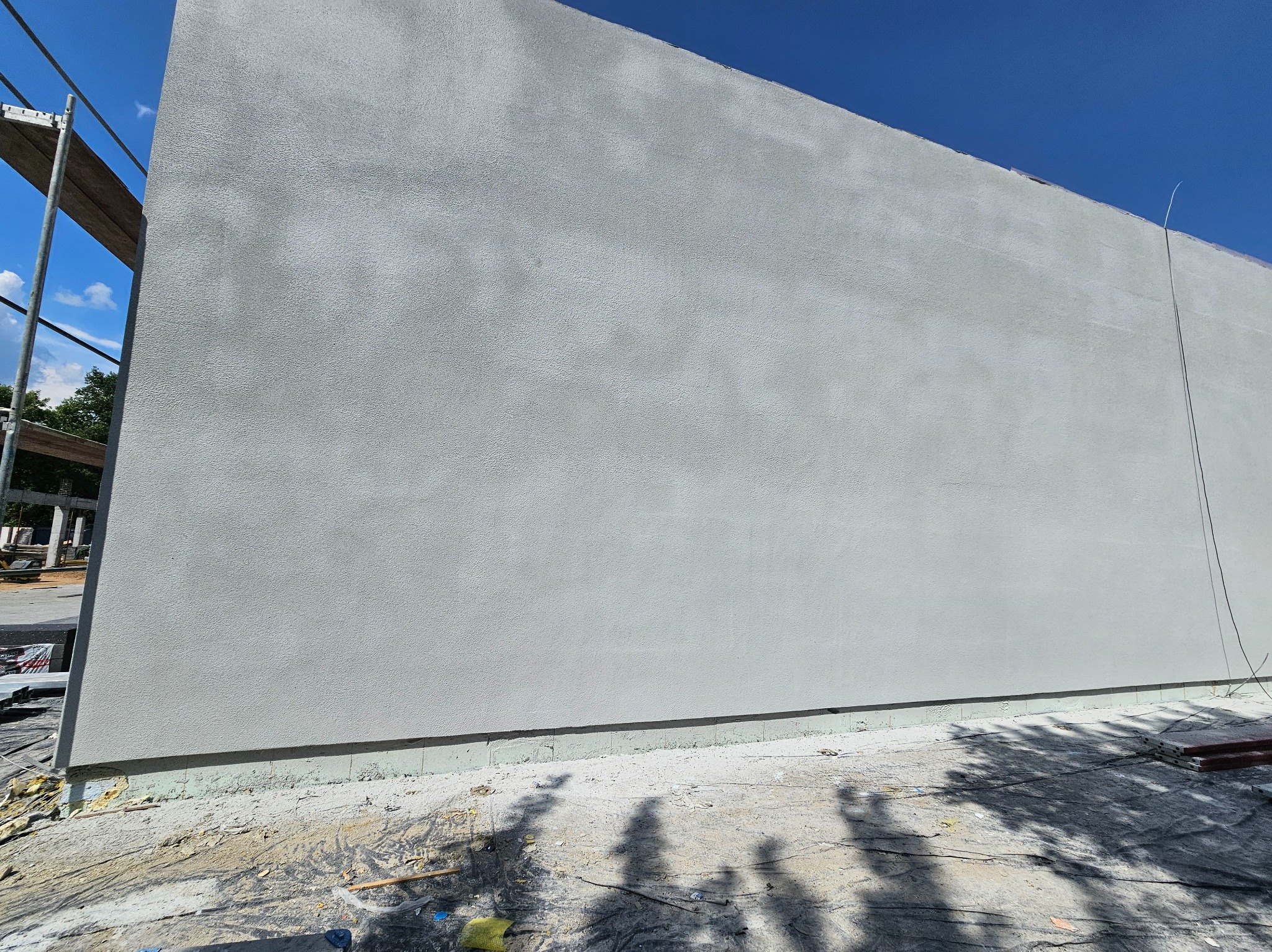 Świeżo otynkowana, gładka ściana budynku w odcieniach szarości, widoczna z dołu pod błękitnym niebem. Fragment rusztowania po lewej stronie. Cień drzewa na dole.