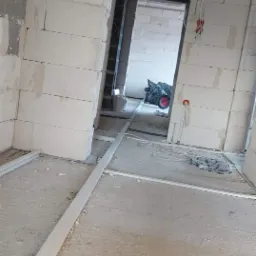 Wnętrze w trakcie prac wykończeniowych: surowe ściany z betonu komórkowego, widoczne przewody elektryczne wychodzące ze ściany, listwy przypodłogowe w trakcie montażu, w tle wózek budowlany.