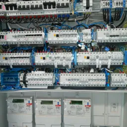 Rozdzielnica elektryczna z bezpiecznikami, przewodami i licznikami energii, widoczne opisy obwodów takie jak 'Hotel 1', 'Gniazda Kuch', 'Pompy', zróżnicowane kolory przewodów, zainstalowane...