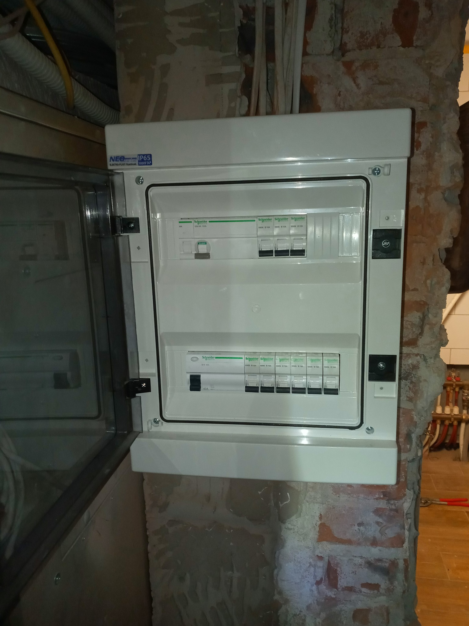 Otwarta skrzynka rozdzielcza Schneider zamontowana na surowej, ceglanej ścianie w trakcie remontu, widoczne kable i rury.