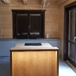 Nowoczesna kuchnia w domu z bali: szare szafki, drewniana wyspa z podświetleniem, czarny zlew pod oknem i przesuwne drzwi balkonowe.