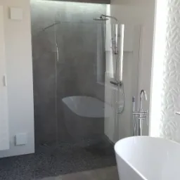 Nowoczesna łazienka z wolnostojącą wanną, kabiną prysznicową typu walk-in z szarą ścianą wykończoną kamieniami oraz białą toaletą.